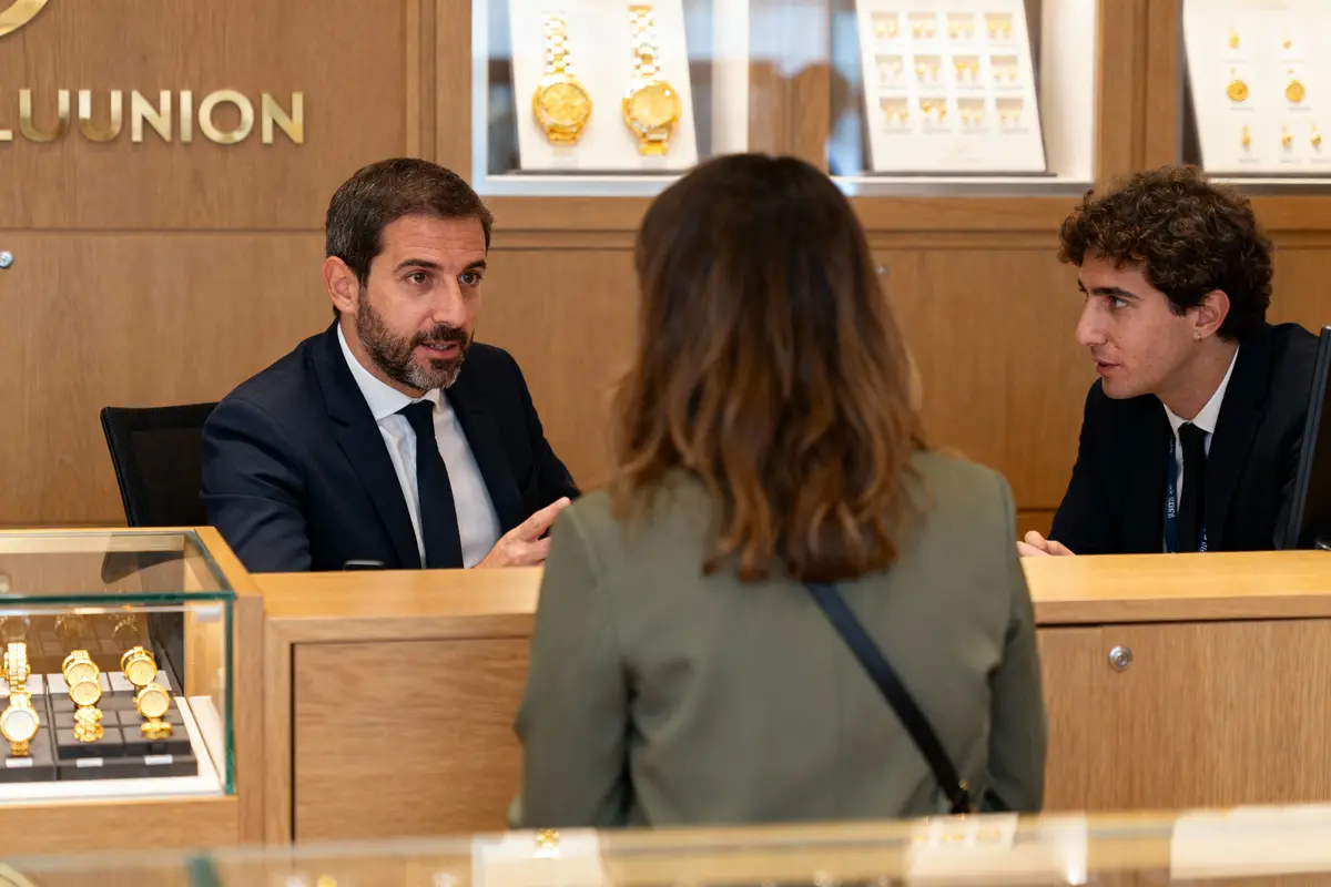 Deux hommes en costume parlent avec une femme dans une bijouterie, montres en arrière-plan.