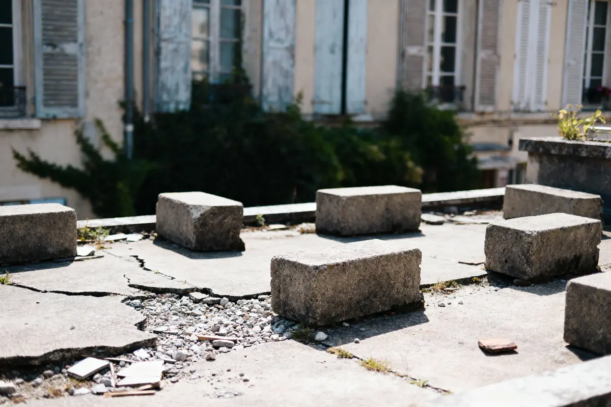 pexels-michal-pech-213601-757318 (1) Blocs de béton sur toit fissuré, devant bâtiment ancien avec volets en bois. Végétation en arrière-plan.