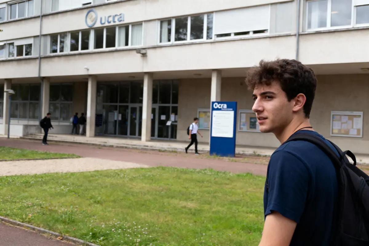 pexels-michal-pech-213601-757318 (1) Un jeune homme devant un bâtiment universitaire avec logo "ucra", portant un sac à dos, d'autres étudiants présents.