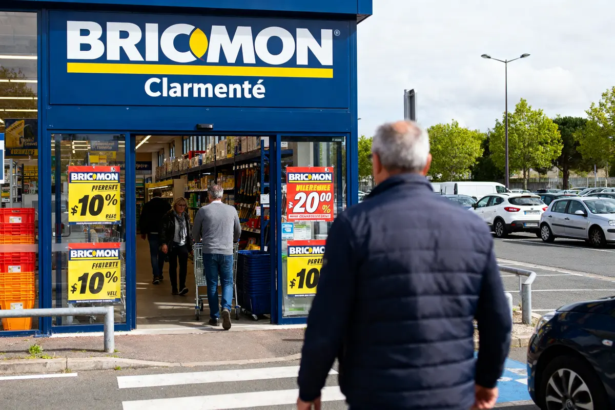 pexels-michal-pech-213601-757318 (1) Entrée d'un magasin Bricomon avec des promotions affichées, clients entrant. Parking avec voitures en arrière-plan.