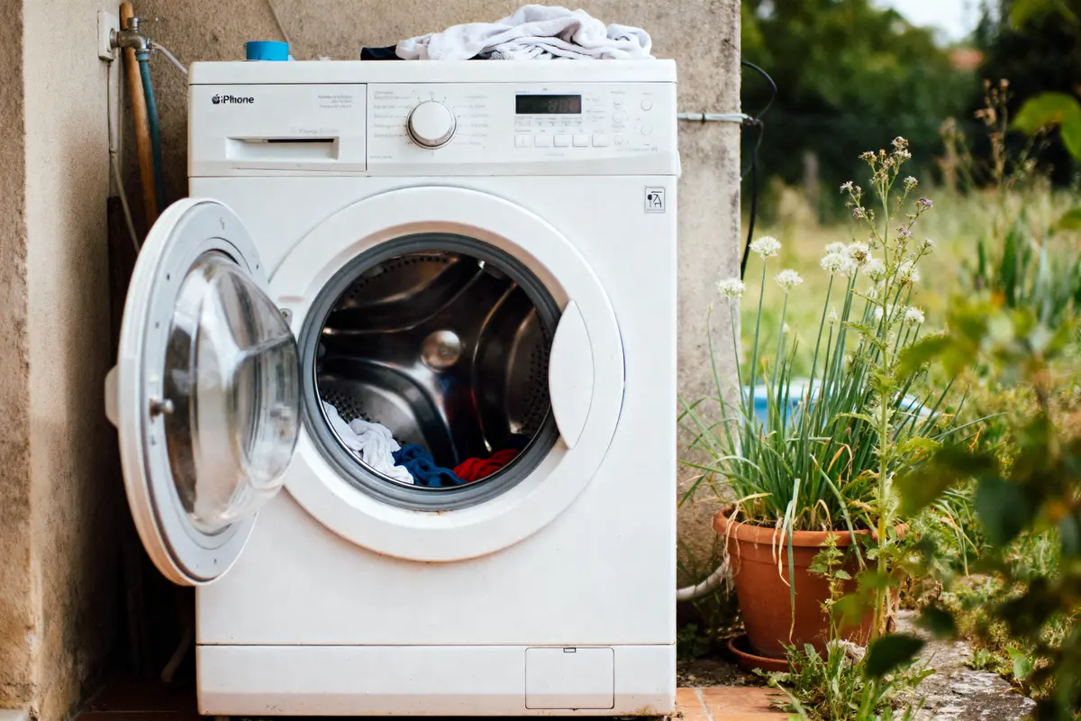 pexels-michal-pech-213601-757318 (1) Machine à laver blanche, porte ouverte, linge à l'intérieur, plante en pot à côté, extérieur verdoyant.