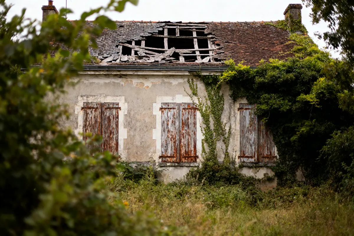 pexels-michal-pech-213601-757318 (1) Maison abandonnée avec toit effondré, volets en bois usés et végétation envahissante.