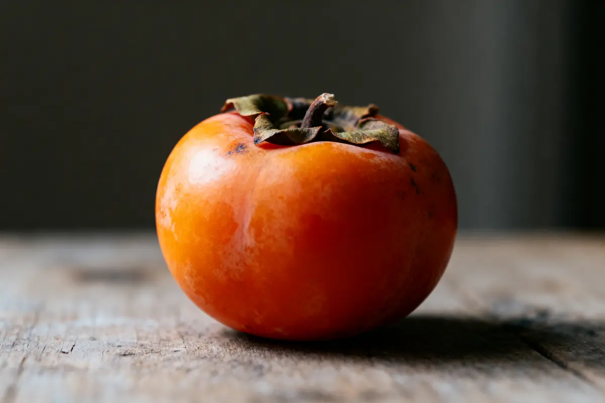 pexels-michal-pech-213601-757318 (1) Kaki orange mûr posé sur une table en bois avec fond sombre flou.